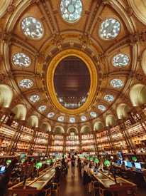 A grand and ornate library interior featuring a large, decorative ceiling with elaborate designs and skylights. Walls are lined with tall bookshelves filled with books, creating a circular pattern around the room. The center is occupied by numerous tables with green lamps, where people are seated reading and working on computers. The atmosphere is lively yet studious.
