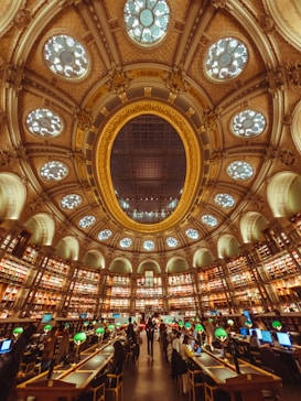A grand and ornate library interior featuring a large, decorative ceiling with elaborate designs and skylights. Walls are lined with tall bookshelves filled with books, creating a circular pattern around the room. The center is occupied by numerous tables with green lamps, where people are seated reading and working on computers. The atmosphere is lively yet studious.