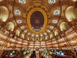 A grand library interior with an ornate, circular design. The ceiling is adorned with an impressive dome featuring decorative patterns and circular light fixtures. Bookshelves line the curved walls, filled with books, and the space is illuminated by green reading lamps. People are seated at desks with computers, engaging in study or work.