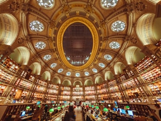 A grand library interior with an ornate, circular design. The ceiling is adorned with an impressive dome featuring decorative patterns and circular light fixtures. Bookshelves line the curved walls, filled with books, and the space is illuminated by green reading lamps. People are seated at desks with computers, engaging in study or work.