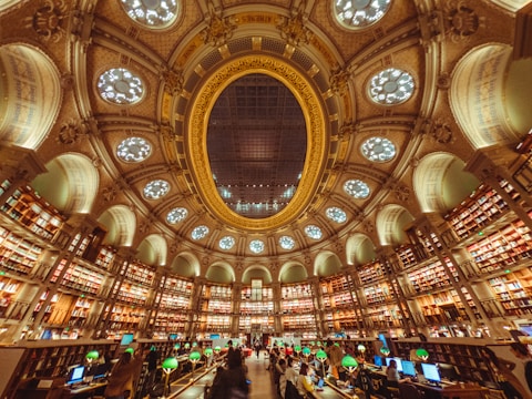 A grand library interior with an ornate, circular design. The ceiling is adorned with an impressive dome featuring decorative patterns and circular light fixtures. Bookshelves line the curved walls, filled with books, and the space is illuminated by green reading lamps. People are seated at desks with computers, engaging in study or work.