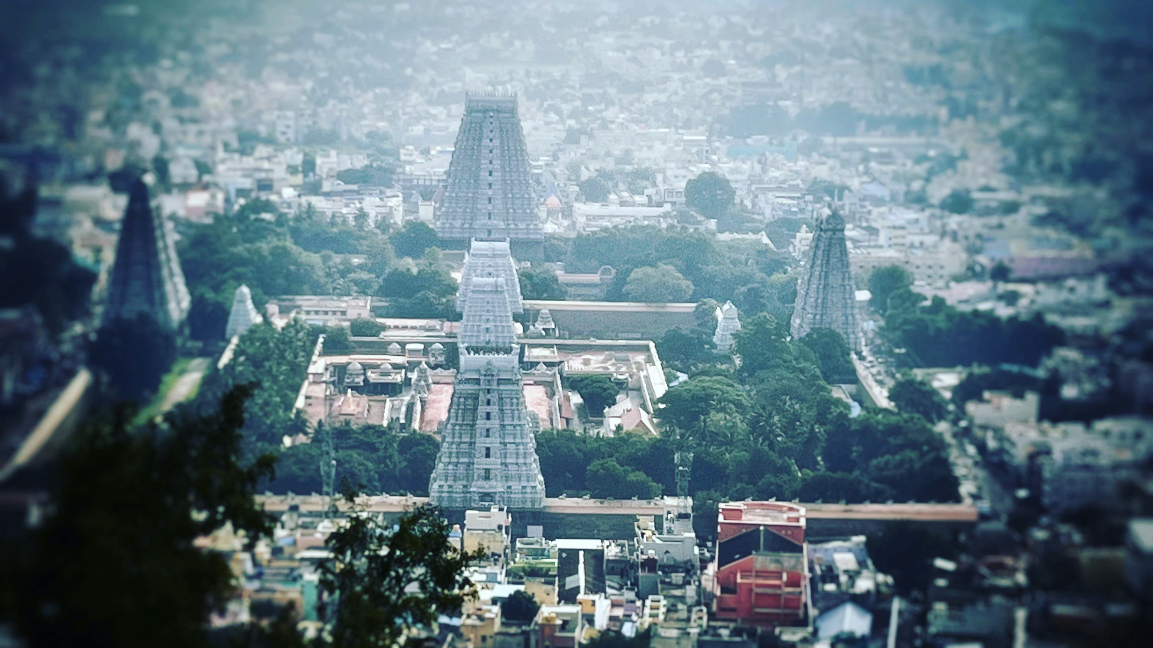 Aerial view of a large temple complex with multiple towering structures, surrounded by greenery and urban sprawl. Architectural towers with intricate designs rise prominently in the center, indicating religious significance.
