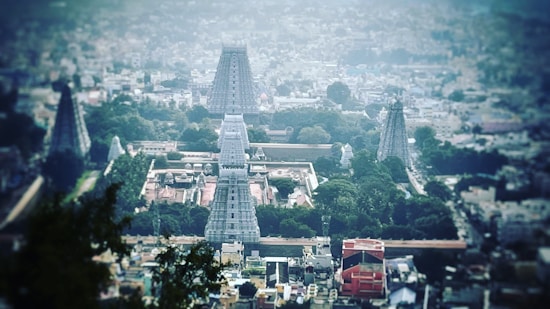Aerial view of a large temple complex with multiple towering structures, surrounded by greenery and urban sprawl. Architectural towers with intricate designs rise prominently in the center, indicating religious significance.