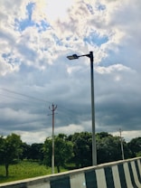 A streetlight on a pole stands alongside a road with a striped barrier. The sky is filled with dense clouds, allowing patches of blue to peek through. In the background, trees and lush greenery stretch out across the landscape, with power lines running parallel to the road.