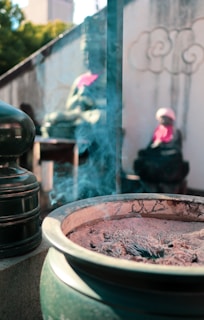 A close-up of a large incense burner filled with ashes and smoldering incense sticks. Bluish smoke rises gently from the ashes, creating an ethereal atmosphere. In the background, two statues adorned with bright pink bibs are visible. The scene is set against a textured wall with cloud patterns.