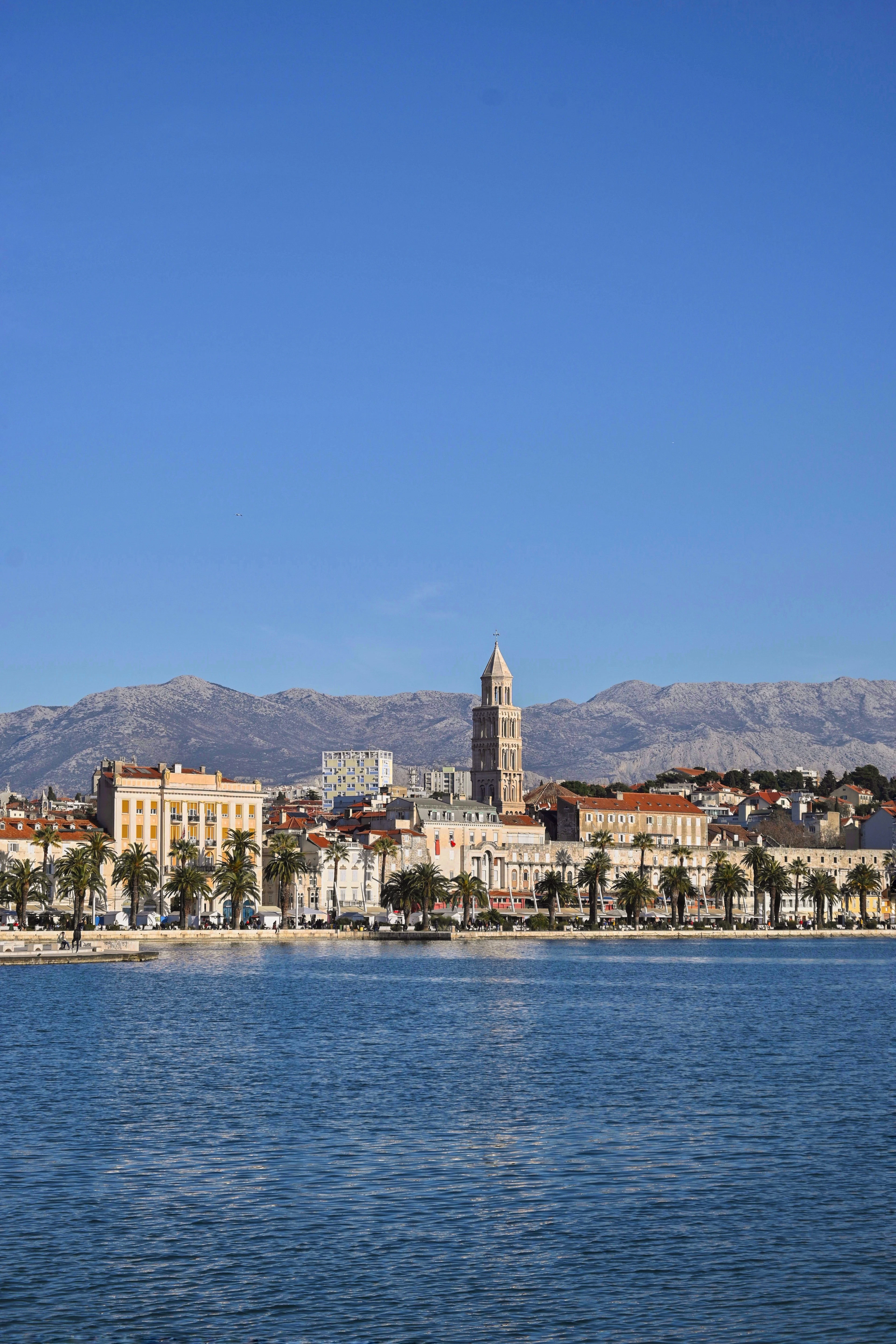 The waterfront of Split seen from out on the Adriatic, the palm-lined Riva running along the cream stone facades of the old town with the grey Dalmatian mountains rising behind under a clear blue sky.