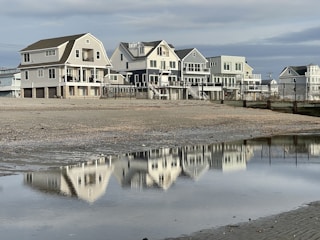 Several coastal houses line a beach with their reflections visible in a large puddle of water in the foreground. The sky is cloudy, creating a serene and calm atmosphere.