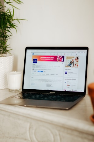 A laptop displaying a LinkedIn profile page is placed on a light-colored surface. To the left of the laptop, there is a white candle in a glass holder and a green plant in a white textured pot.