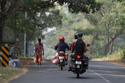 A rural road with two motorcyclists wearing helmets and carrying backpacks riding away from the camera. A woman is walking on the side of the road carrying bags and a bottle, surrounded by lush green trees.