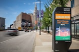 An urban street scene in Cleveland with a prominent digital kiosk displaying a message about clean energy, jobs, and justice. The background features tall buildings, including a distinctive historic skyscraper, and a large billboard with 'For the Love of Land' visible. Vehicles are visible on the road, some in motion creating a blur effect, while trees line the sidewalk.