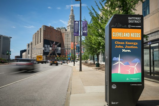 An urban street scene in Cleveland with a prominent digital kiosk displaying a message about clean energy, jobs, and justice. The background features tall buildings, including a distinctive historic skyscraper, and a large billboard with 'For the Love of Land' visible. Vehicles are visible on the road, some in motion creating a blur effect, while trees line the sidewalk.