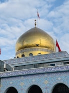 A grand structure featuring a large golden dome adorned with Islamic architectural elements. The dome is topped with intricate ornamentation and flanked by red flags. The building's facade is decorated with detailed blue and white mosaic tiles, featuring Arabic script and geometric patterns. A bird is captured in flight near the dome.