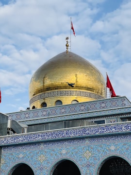 A grand structure featuring a large golden dome adorned with Islamic architectural elements. The dome is topped with intricate ornamentation and flanked by red flags. The building's facade is decorated with detailed blue and white mosaic tiles, featuring Arabic script and geometric patterns. A bird is captured in flight near the dome.