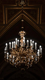 A grand, ornate chandelier with multiple candle-like bulbs hanging in a lavish setting. The fixture is intricately designed with gold and bronze elements, casting a warm glow in the dimly lit room. The ceiling features decorative patterns and a triangular arch framing the chandelier.