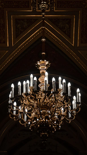 A grand, ornate chandelier with multiple candle-like bulbs hanging in a lavish setting. The fixture is intricately designed with gold and bronze elements, casting a warm glow in the dimly lit room. The ceiling features decorative patterns and a triangular arch framing the chandelier.