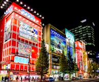 A vibrant city scene at night featuring large illuminated buildings lined with colorful and bright advertisements. Prominent signage for gaming and electronics companies such as SEGA can be seen, and the streets are bustling with pedestrians and cars. Tall trees line the street, adding a touch of greenery to the urban setting.