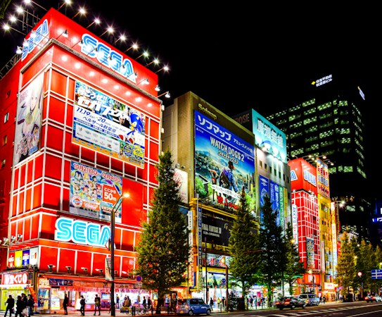A vibrant city scene at night featuring large illuminated buildings lined with colorful and bright advertisements. Prominent signage for gaming and electronics companies such as SEGA can be seen, and the streets are bustling with pedestrians and cars. Tall trees line the street, adding a touch of greenery to the urban setting.