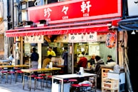 A street-side restaurant features a vibrant red awning with Japanese characters. Outdoor tables and chairs are arranged neatly, and several people are seated, enjoying their meals. Inside, a person in a yellow shirt is visible, likely preparing food. The atmosphere appears lively and casual, typical of an urban dining setting.