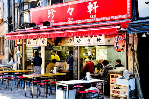 A street-side restaurant features a vibrant red awning with Japanese characters. Outdoor tables and chairs are arranged neatly, and several people are seated, enjoying their meals. Inside, a person in a yellow shirt is visible, likely preparing food. The atmosphere appears lively and casual, typical of an urban dining setting.