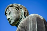 A large, detailed bronze statue of Buddha with tightly curled hair and closed eyes against a clear blue sky. The statue's serene expression and gentle contours suggest tranquility and wisdom.