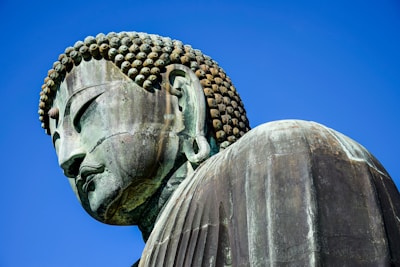 A large, detailed bronze statue of Buddha with tightly curled hair and closed eyes against a clear blue sky. The statue's serene expression and gentle contours suggest tranquility and wisdom.