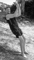 A person is enjoying an outdoor activity, holding onto a rope while balancing on their feet. The individual is dressed in athletic clothing and appears to be smiling, indicating they are experiencing joy or excitement. The surroundings are natural, with trees and leaves scattered on the ground, suggesting a forest or park setting.