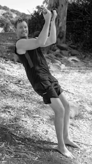 A person is enjoying an outdoor activity, holding onto a rope while balancing on their feet. The individual is dressed in athletic clothing and appears to be smiling, indicating they are experiencing joy or excitement. The surroundings are natural, with trees and leaves scattered on the ground, suggesting a forest or park setting.