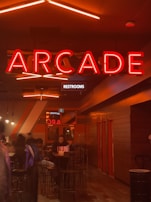 A dimly lit interior features a large red neon sign reading 'ARCADE' above a long hallway with tables and chairs. The atmosphere is dark, with a warm glow from the lighting fixtures and walls. Several people are seated at tables in a social setting, creating a lively but cozy ambiance.