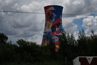 A large cooling tower is vividly painted with colorful murals featuring diverse human figures, hands, and various symbols. The tower stands amidst lush green trees under a cloudy sky.
