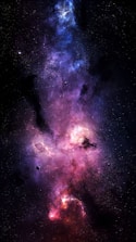 Vivid hues of purples, blues, and pinks blend seamlessly with specks of white representing distant stars against a backdrop of deep space. A celestial cloud of gas and dust forms a nebula, illuminated by the light of nearby stars. Amidst the vibrant colors, a small, dark planet orbits in the foreground.