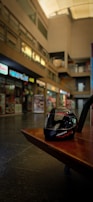 A motorcycle helmet rests on a brown wooden bench in an indoor shopping area with various storefronts displaying signs and menus. The background is blurred, highlighting the helmet in the foreground.