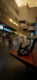 A motorcycle helmet rests on a brown wooden bench in an indoor shopping area with various storefronts displaying signs and menus. The background is blurred, highlighting the helmet in the foreground.