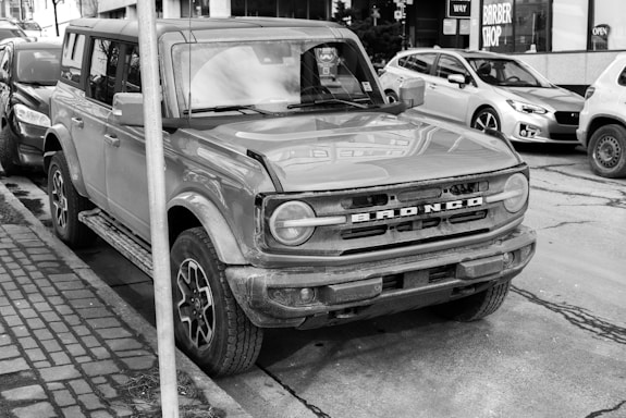 A rugged SUV is parked on a city street, partially obstructed by a pole. It has a boxy design with noticeable dirt on the body, suggesting it has been used for off-road or in tough conditions. Other cars are parked nearby, with a barbershop visible in the background.