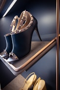 A pair of elegant black high-heeled shoes adorned with gold studded straps displayed on a shelf. The shoes are positioned on an angled rack with another pair of beige shoes visible on a lower shelf. The lighting and shelves give a luxurious and sophisticated ambiance.