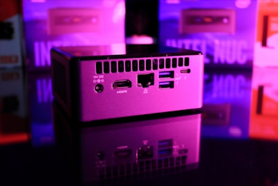 A compact computer unit sits on a reflective surface, surrounded by colorful lighting. The front panel features various ports including HDMI and USB, and bright pink and purple hues dominate the background, creating a futuristic atmosphere.