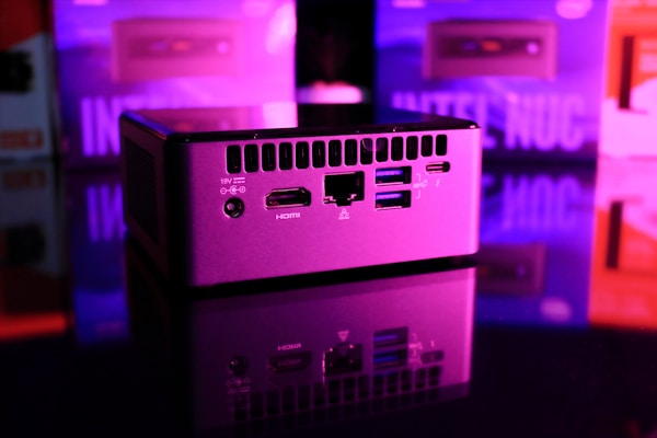 A compact computer unit sits on a reflective surface, surrounded by colorful lighting. The front panel features various ports including HDMI and USB, and bright pink and purple hues dominate the background, creating a futuristic atmosphere.