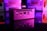 A compact computer unit sits on a reflective surface, surrounded by colorful lighting. The front panel features various ports including HDMI and USB, and bright pink and purple hues dominate the background, creating a futuristic atmosphere.