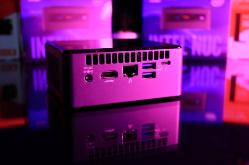 A compact computer unit sits on a reflective surface, surrounded by colorful lighting. The front panel features various ports including HDMI and USB, and bright pink and purple hues dominate the background, creating a futuristic atmosphere.