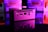 A compact computer unit sits on a reflective surface, surrounded by colorful lighting. The front panel features various ports including HDMI and USB, and bright pink and purple hues dominate the background, creating a futuristic atmosphere.