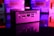 A compact computer unit sits on a reflective surface, surrounded by colorful lighting. The front panel features various ports including HDMI and USB, and bright pink and purple hues dominate the background, creating a futuristic atmosphere.