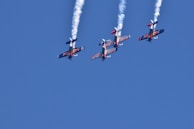 Four aerobatic airplanes fly closely together in an inverted formation, leaving white smoke trails against a clear blue sky. The planes display vibrant colors and branding on their wings.