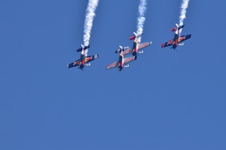 Four aerobatic airplanes fly closely together in an inverted formation, leaving white smoke trails against a clear blue sky. The planes display vibrant colors and branding on their wings.