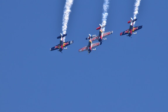 Four aerobatic airplanes fly closely together in an inverted formation, leaving white smoke trails against a clear blue sky. The planes display vibrant colors and branding on their wings.