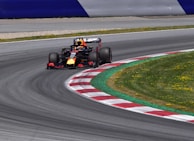 A Formula 1 race car speeds through a curved section of a race track. The track features red and white kerbs alongside a well-maintained grassy area. The car is adorned with vibrant colors and sponsors' logos. The setting is bright, indicating a clear day.