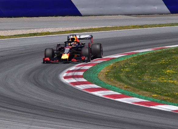 A Formula 1 race car speeds through a curved section of a race track. The track features red and white kerbs alongside a well-maintained grassy area. The car is adorned with vibrant colors and sponsors' logos. The setting is bright, indicating a clear day.