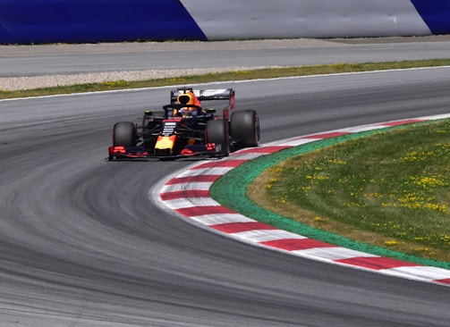 A Formula 1 race car speeds through a curved section of a race track. The track features red and white kerbs alongside a well-maintained grassy area. The car is adorned with vibrant colors and sponsors' logos. The setting is bright, indicating a clear day.