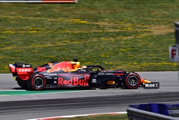 A Red Bull Formula 1 racing car is captured in motion on a track, featuring prominent branding and logos from various sponsors. The car's sleek design and speed are emphasized by its low profile and aerodynamic shape. In the background, a grassy area is visible with patches of yellow flowers.