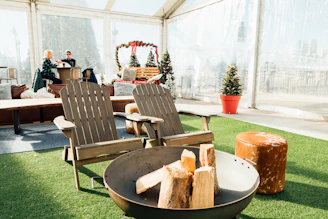 Two wooden Adirondack chairs face a round fire pit filled with logs, suggesting an outdoor lounging area. Behind them, two people are sitting at a bar-like counter, possibly enjoying a drink. The setting is festive, with small decorated Christmas trees placed around the area. The space is covered by a transparent tent and the floor is laid with artificial grass.