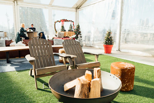 Two wooden Adirondack chairs face a round fire pit filled with logs, suggesting an outdoor lounging area. Behind them, two people are sitting at a bar-like counter, possibly enjoying a drink. The setting is festive, with small decorated Christmas trees placed around the area. The space is covered by a transparent tent and the floor is laid with artificial grass.