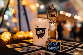 A gold skeleton trophy labeled 'Best Group' is prominently displayed on a table. Next to it, there is a white candle with the label 'Boos & Brews on the Lake.' The background features blurred, warm-toned bokeh lights adding a festive, cozy atmosphere. Several Polaroid photos are scattered on the table.